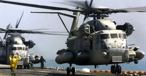 A military helicopter hovers over a ship deck with personnel nearby.