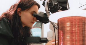 A woman closely examines a copper coil under a microscope in a lab.